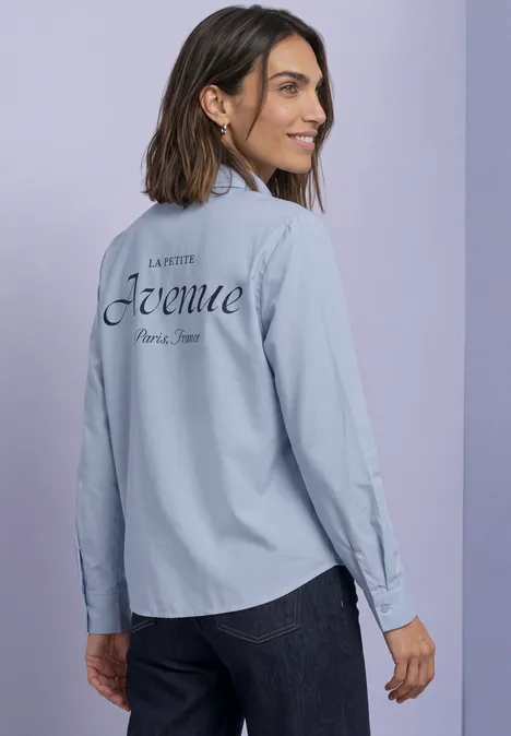 Frau in hellblauem Hemd mit Aufdruck 'Avenue Paris New' auf dem Rücken, Blick über die Schulter, lächelnd.
