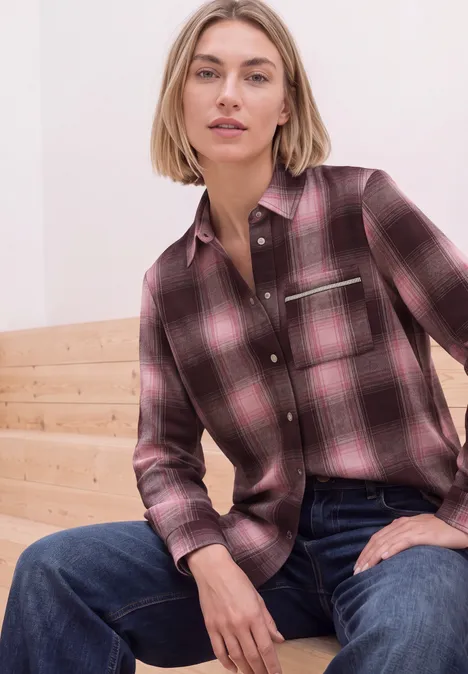 Frau in rosa kariertem Hemd und blauen Jeans sitzt entspannt auf hellen Holzstufen.
