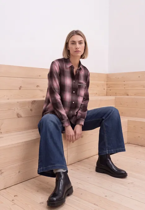 Frau in karierter Bluse, weitem Jeans und schwarzen Stiefeletten sitzt auf Holzbänken.
