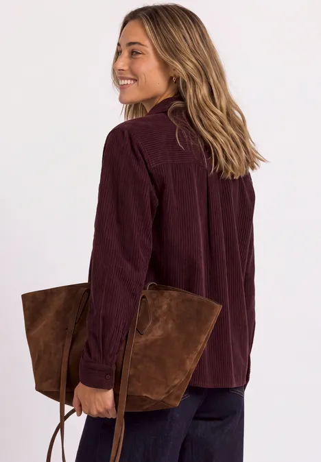 Frau in bordeauxfarbenem Cordhemd, lächelnd, hält große braune Wildledertasche.