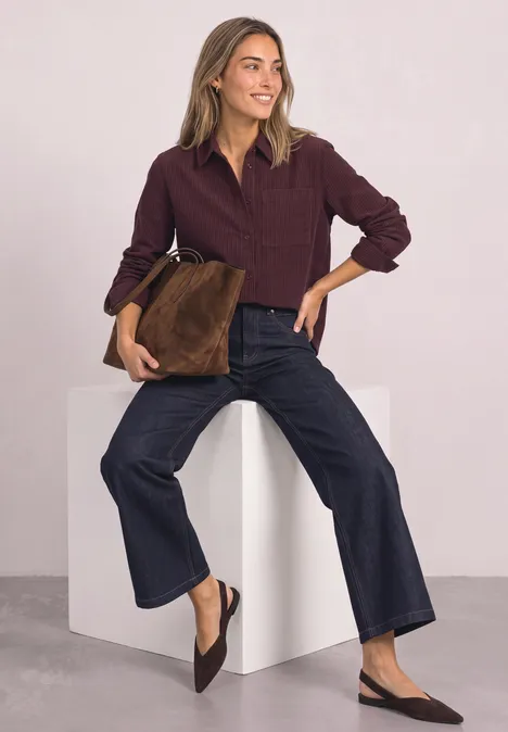 Frau in kastiger Cordbluse, weiten Jeans und Slingbacks, hält eine braune Veloursleder-Tasche vor hellem Hintergrund.