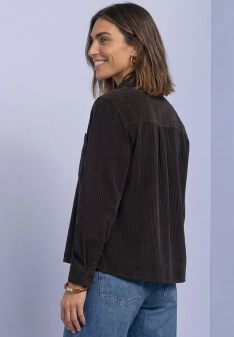 Frau in brauner Cordjacke und blauer Jeans vor pastellfarbenem Hintergrund lächelt seitlich