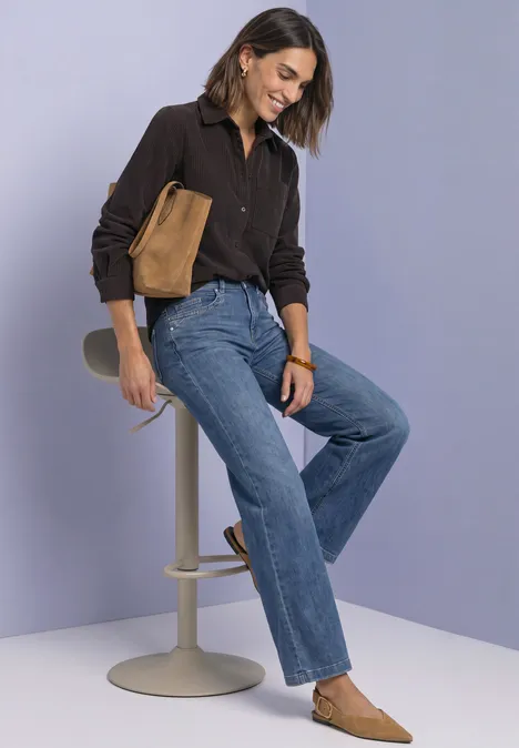 Frau in casual Outfit, Jeans, braunen Schuhen und passender Tasche sitzt entspannt auf Hocker vor hellem Hintergrund.
