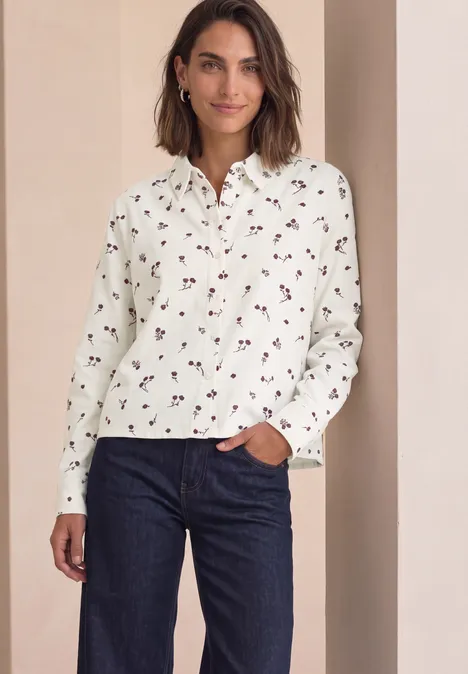 Eine Frau trägt eine weiße Bluse mit kleinem Blumenmuster und dunkelblaue Jeans vor neutralem Hintergrund.