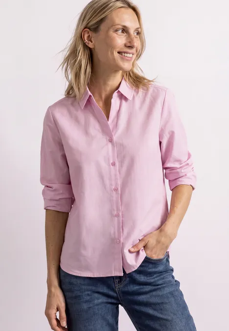 Eine lächelnde Frau mit rosa Hemd und Jeans vor hellem Hintergrund.