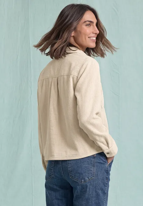Een lachende vrouw met halflang donker haar, draagt een beige jasje en spijkerbroek, zijaanzicht voor een lichte achtergrond.