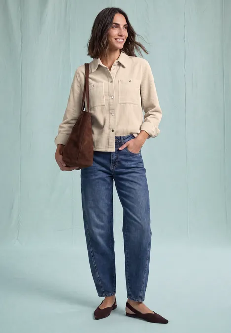 Een glimlachende vrouw in een beige jasje, blauwe jeans, bruine schoenen en schoudertas.
