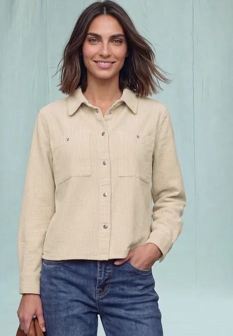 Een vrouw draagt een beige overhemd en blauwe jeans, staat voor een lichte achtergrond en lacht.