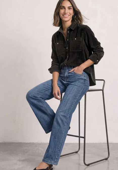 Overhemd van corduroy met borstzakken black coffee Frau auf hohem Stuhl in schwarzer Bluse und blauen Jeans, sie sitzt entspannt und lächelt.