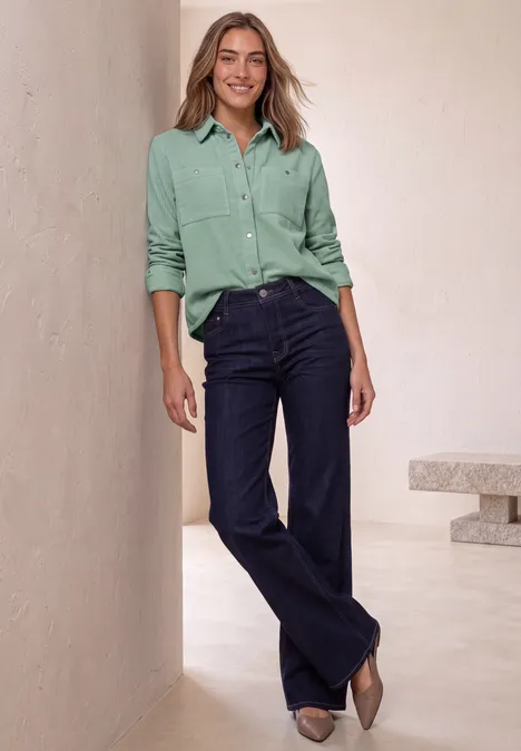 Overhemdblouse van corduroy met borstzakken eucalyptus green Eine lächelnde Frau lehnt an einer Wand, trägt eine mintgrüne Bluse, weite dunkelblaue Jeans und beigen Pumps.