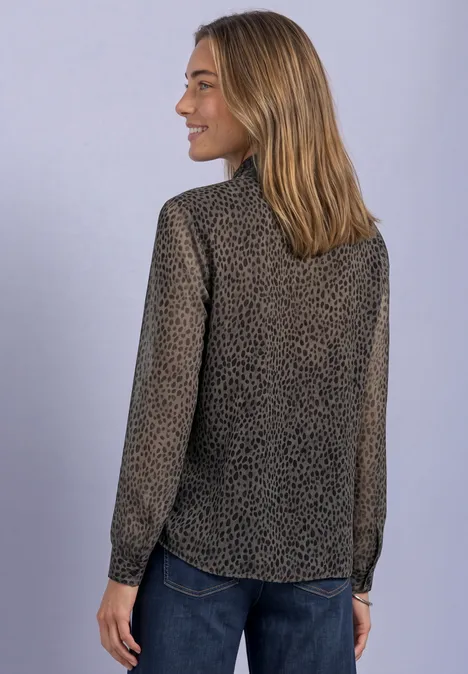 Eine Frau in Profilansicht trägt eine gepunktete Bluse mit langen Ärmeln und Jeans vor grauem Hintergrund.