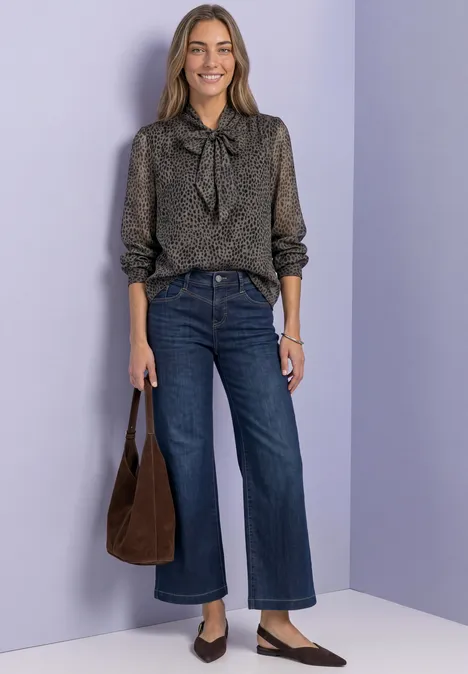 Frau in geblümter Schlupfbluse mit Schleife, weiten Jeans, braune Tasche und spitzen Loafers vor lila Hintergrund.