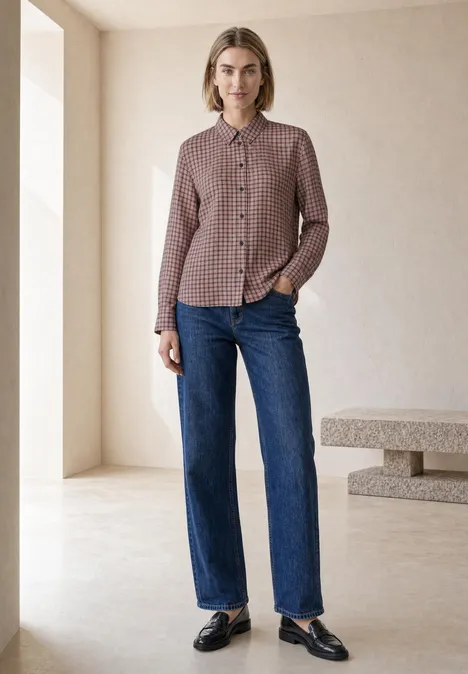 Eine Frau steht in minimalistischem Raum, trägt karierte Bluse, gerade geschnittene Jeans und schwarze Loafers.