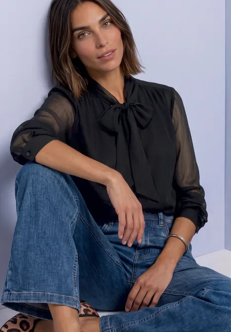 Eine Frau in schwarzer Bluse mit Schleife und blauen Jeans sitzt vor hellem Hintergrund.