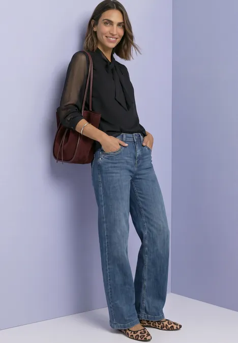 Frau in schwarzer Bluse mit Bindedetail, blauen Jeans, Leo-Pumps und Schultertasche vor lila Hintergrund.