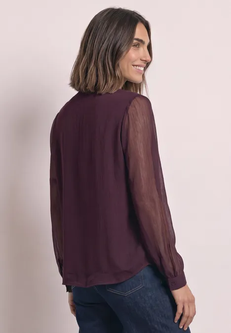 Frau steht seitlich, lächelt, trägt eine dunkle, transparente Bluse mit langen Ärmeln und dunkle Jeans.