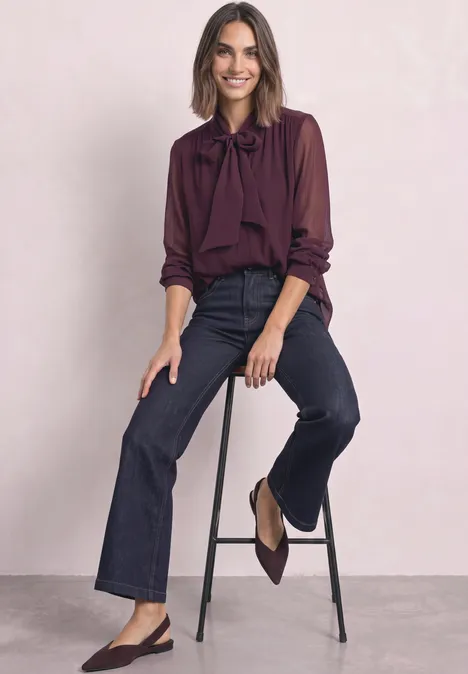 Dame sitzt auf Hocker, trägt weinrote Bluse mit Schleife, dunkelblaue Jeans und Slingback-Pumps.