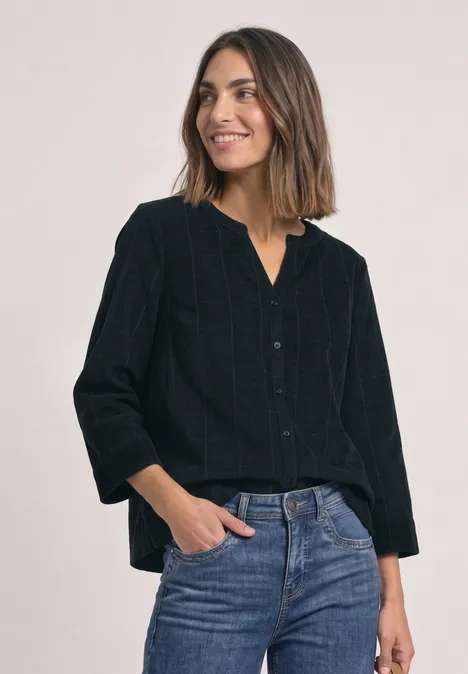 Frau lächelt, trägt schwarze Bluse und Jeans, steht entspannt mit Händen in den Taschen.