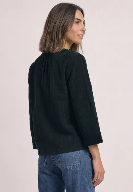 Frau steht seitlich abgewandt, trägt schwarze Bluse mit Struktur und blaue Jeans, schaut über die Schulter.