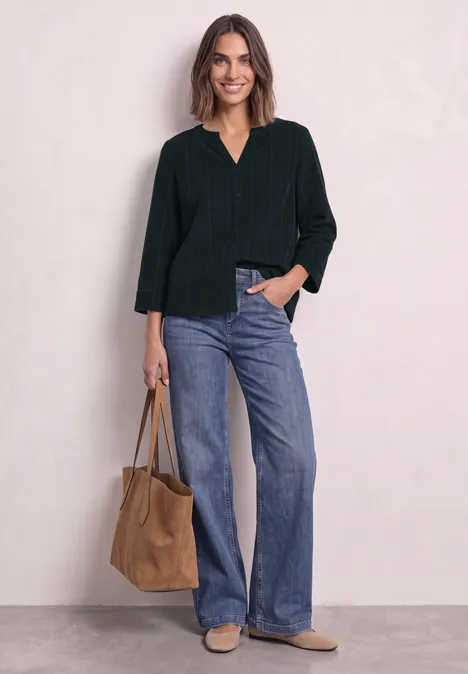 Frau in dunkelgrünem Oberteil und blauer Jeans, lächelnd, hält braune Tasche.