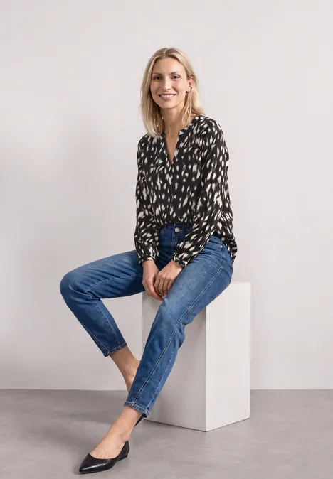 Femme en chemisier noir à motifs et jeans bleus assise élégamment sur un cube blanc.