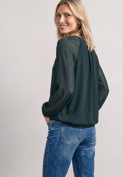 Een glimlachende vrouw in spijkerbroek en donkergroene blouse kijkt over haar schouder in de camera.