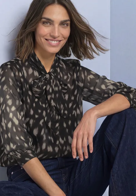 Chemisier à manches longues en chiffon avec nœud décoratif Black Eine lächelnde Frau in gemusterter Bluse und Jeans lehnt entspannt an einer Wand.