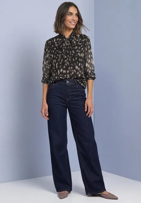 Chemisier à manches longues en chiffon avec nœud décoratif Black Frau trägt geblümte Bluse, dunkelblaue Schlaghose und spitze Schuhe, steht vor hellem lila Hintergrund.