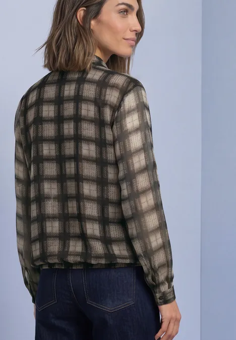 Vrouw draagt halftransparant geruit langarmshirt in zwart-beige, staand opzij met een blik over haar schouder.
