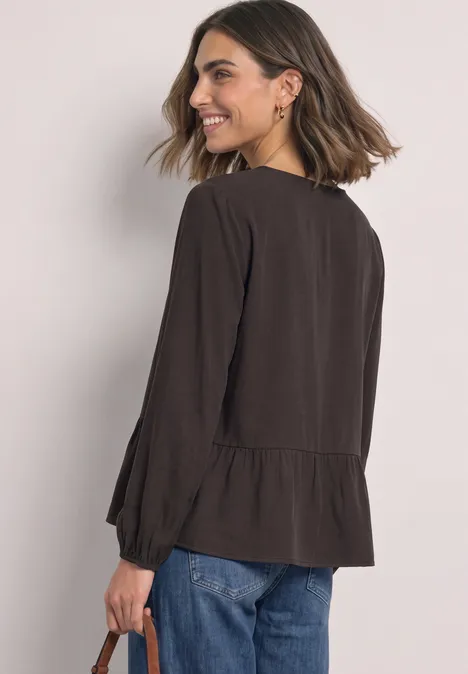 Chemisier à manches longues et à basque black coffee Frau mit schulterlangem Haar trägt schwarze Bluse mit Volant und blaue Jeans, lächelt über die Schulter.