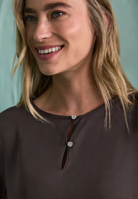 Chemisier à manches longues et à basque black coffee Nahaufnahme einer lächelnden Frau mit blondem Haar, braunem Oberteil und Knöpfen am Ausschnitt.