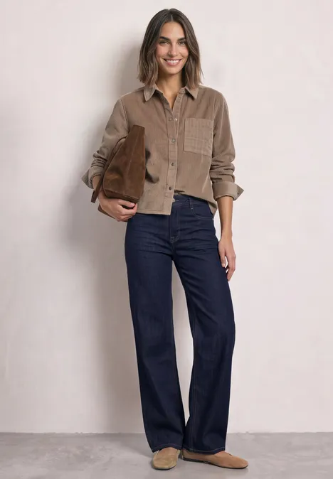 Eine Frau trägt ein braunes Cordhemd, weite Jeans und Hausschuhe, dazu eine braune Tasche.