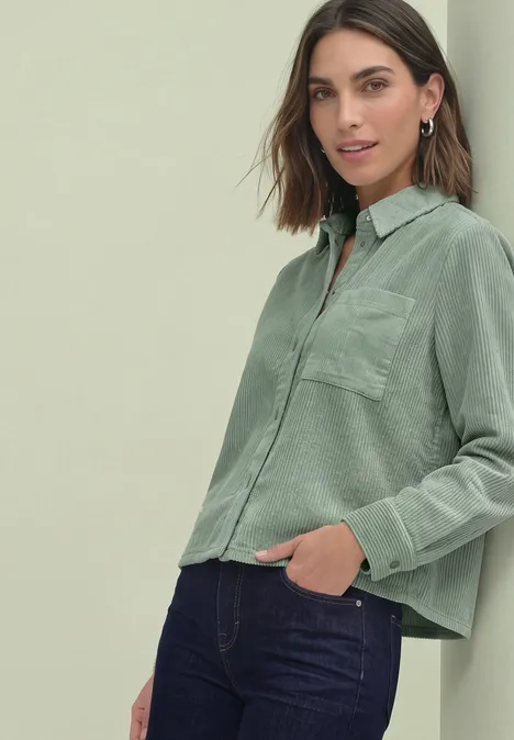 Frau trägt hellgrüne, locker sitzende Bluse mit Brusttasche und dunkle Jeans.