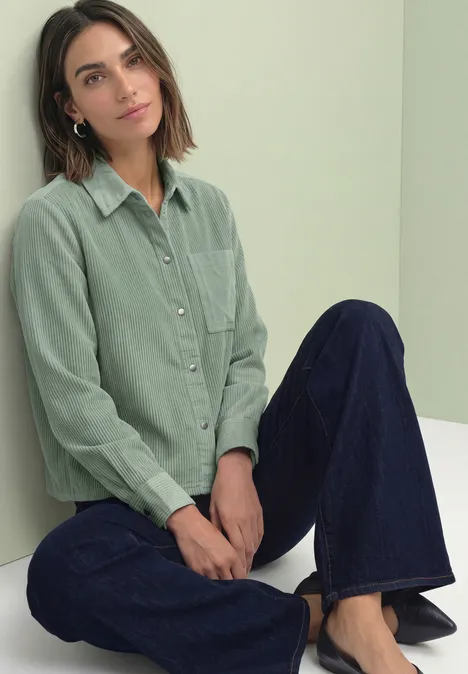 Junge Frau sitzt auf weißem Podest, trägt hellgrünes Cordhemd und weite dunkelblaue Jeans.