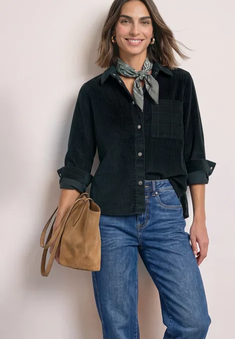 Vrouw met zwarte blouse, blauwe spijkerbroek en bruine schoudertas, met een patroon sjaal, glimlachend.