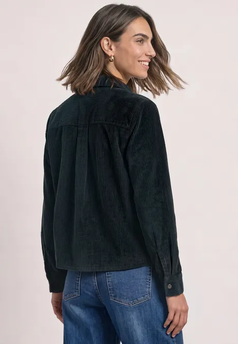 Vrouw in een zwarte corduroyjas en blauwe jeans kijkt lachend over haar schouder.