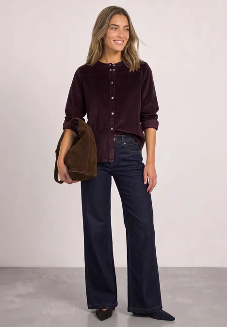Frau im violetten Cordhemd und weiten Jeans, lächelnd, mit brauner Jacke in der Hand.