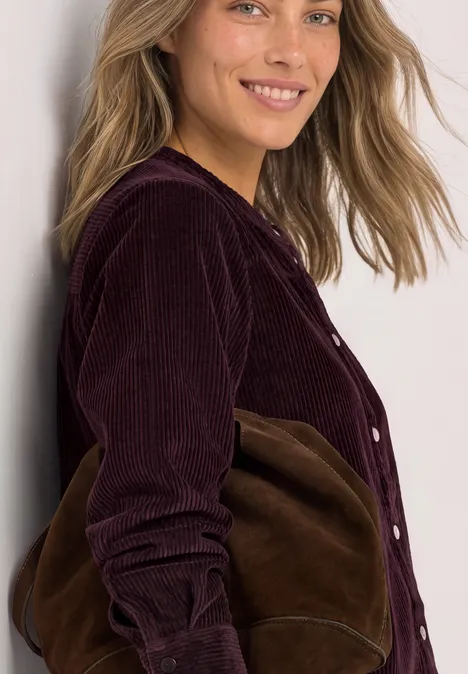 Eine lächelnde Frau lehnt an einer Wand, trägt eine dunkelrote Cordjacke und eine braune Tasche.