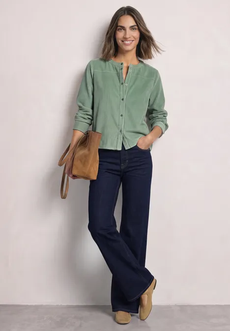 Chemisier à manches longues en velours côtelé avec coutures décoratives eucalyptus green Eine Frau trägt ein grünes Cordhemd, dunkle Schlagjeans, beige Stiefeletten und hält eine Tasche vor hellem Hintergrund.