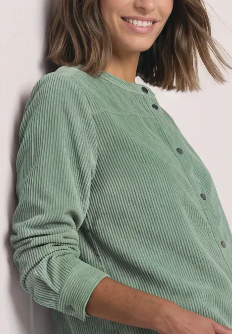 Chemisier à manches longues en velours côtelé avec coutures décoratives eucalyptus green Frau lehnt an weißer Wand, trägt hellgrüne Cordjacke, lächelt leicht.