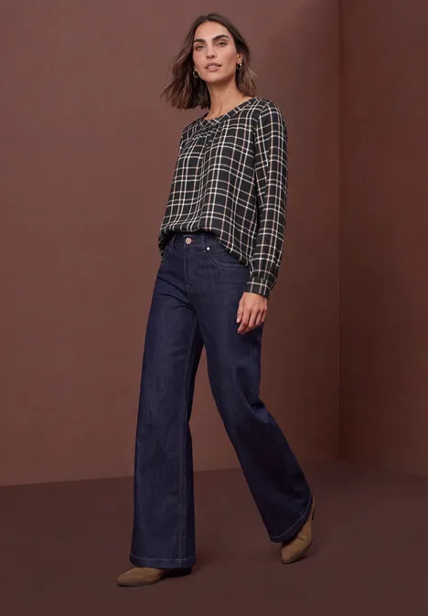 Frau in kariertem Oberteil und weiter High-Waist-Jeans vor braunem Hintergrund.