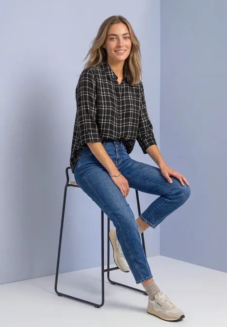 3/4-Arm Hemdbluse mit Karomuster Black Frau sitzt lächelnd auf einem hohen Hocker, trägt kariertes Hemd, Jeans und weiße Sneaker vor schlichter Kulisse.