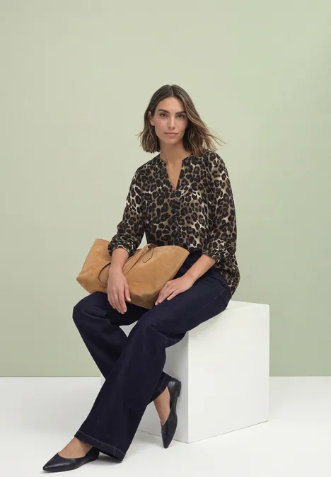 Blouse met lange mouwen, splitnek en luipaardprint Black Vrouw zittend voor lichtgroene achtergrond op witte kubus, luipaardblouse, donkere broek en beige tas.