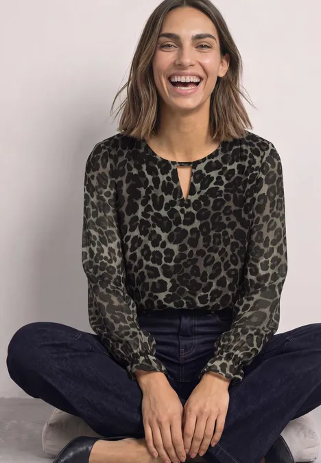Tunika aus Chiffon mit Cut-Out-Detail Black Lachende Frau sitzt crossbeinig in Leoparden-Print-Bluse, dunklen Jeans und Sneakers vor neutralem Hintergrund.
