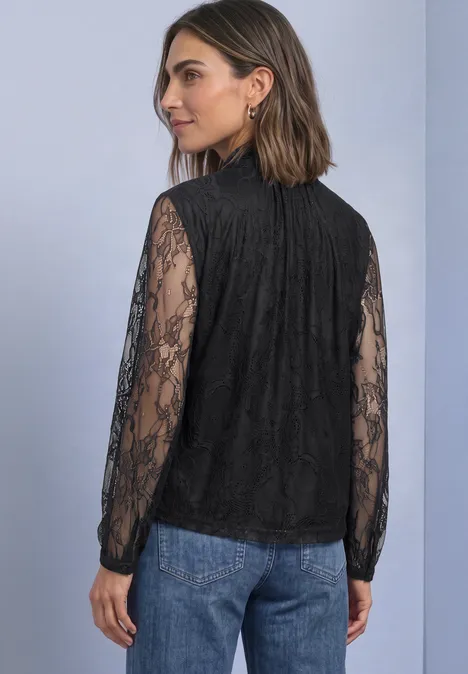 Frau von hinten in schwarzer Spitzenbluse mit langen Ärmeln und blauen Jeans vor neutralem Hintergrund.
