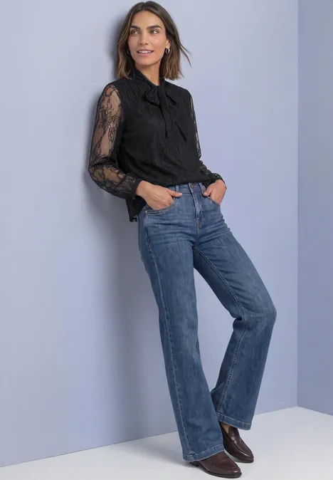 Frau in schwarzer Spitzenbluse und weiten Jeans lehnt an heller Wand und trägt Stiefel.