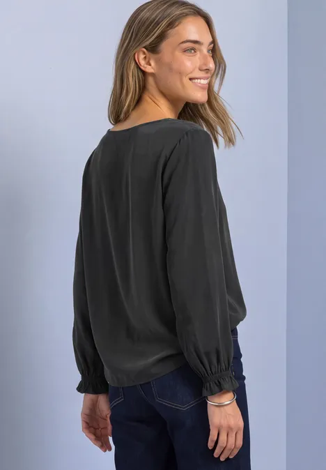 Blouse col rond à boucle décorative armor grey Femme souriante regardant par-dessus son épaule, vêtue d'un chemisier noir à manches longues et d'un jean.