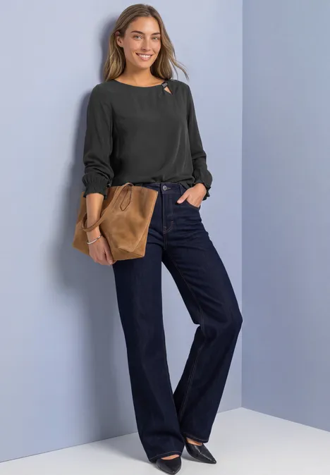 Blouse col rond à boucle décorative armor grey Une femme souriante en pull noir, jean ample et pochette marron, posant devant un mur clair.