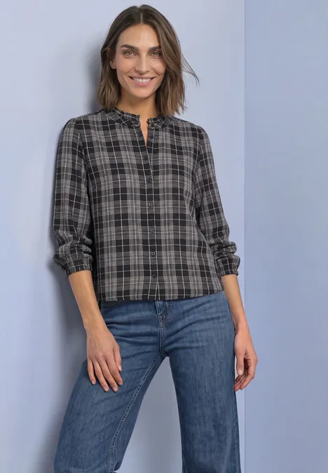 Frau in kariertem Hemd mit Knopfleiste und Jeans vor neutralem Hintergrund.