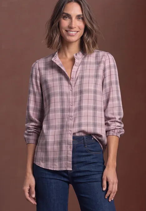 Frau mit rosa kariertem Hemd und Jeans lächelt vor braunem Hintergrund.